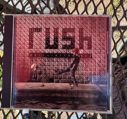 The image features a CD cover for the album "Roll the Bones" by the band Rush. The cover displays a young person kicking a small object in front of a wall made of square, patterned blocks. The overall aesthetic has a reddish hue.