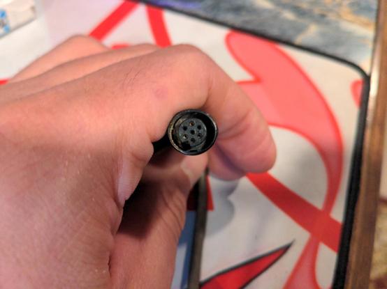 A small round plug, held in the photographer's hand. The plug features seven pins, six arranged around a seventh central pin similar to the cylinder of a revolver. The plug's purpose is a mystery.