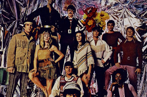 1970 A Band of Counterculture Musicians in a Colorful Photo Spread

The image depicts an eclectic group of individuals, likely musicians or artists from the 1970s, dressed in distinctive and vibrant attire.
They are posed against a backdrop with silver metallic foil which adds to their unconventional appearance.