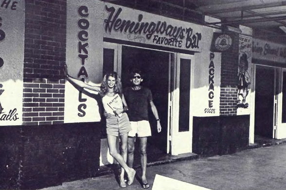1976 

The image shows a couple posing in front of the entrance to Hemingway's Bar, as indicated by signage above them.
They are standing on what appears to be a sidewalk or pavement outside a building.
Tags: black-and-white photograph, vintage photo, 1970s fashion, urban street scene, bar sign