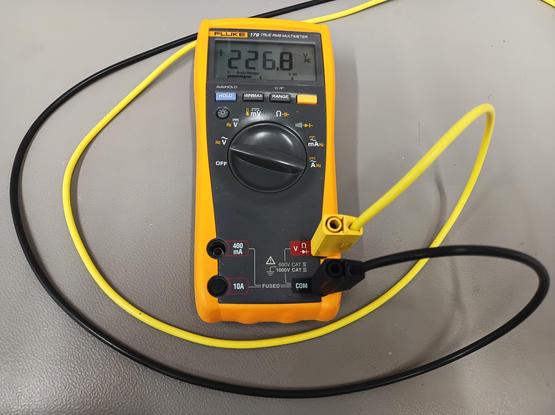 Ein Digitalmultimeter vom Typ Fluke 179 liegt auf einem Labortisch mit grauer ESD-Unterlage. Eine gelbe Messleitung ist an den Eingang für "V" angeschlossen, eine schwarze Messleitung an "COM". Die Messleitungen führen erst um das Gerät herum und dann nach oben rechts aus dem Bild.

Das Gerät steht in Position "V AC" und zeigt "226,8 V AC" auf dem Display.

Seine Farbgebung ist typisch in rutschfester, gelber Gummierung mit grauer Front.
