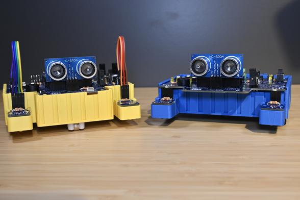 Front view of two 3D-printed BEAPER Bot robots controlled by Arduino Nano ESP32 microcontrollers mounted in BEAPER Nano circuit boards. The front of the yellow robot (left) shows its two optical floor sensor modules connected by extension cables and a centrally mounted ultrasonic SONAR distance sensor module on top of the robot with a ball castor underneath. The front of the blue robot (right) shows its two optical sensor modules connected under the main PCB which also has an ultrasonic SONAR distance sensor module mounted on top.
