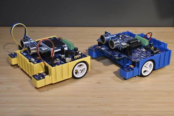 Side view of two 3D-printed BEAPER Bot robots controlled by Arduino Nano ESP32 microcontrollers installed in beginner BEAPER Nano circuit boards. The yellow robot (left) is set up in a rear-drive configuration and includes two optical sensor modules connected by short extension cables, an ultrasonic SONAR distance sensor module, and has a small graphical LCD panel installed on top. The blue robot (right) is set up in a front-drive configuration and includes two optical sensor modules connected by headers under the PCB and an ultrasonic SONAR distance sensor module installed on top.