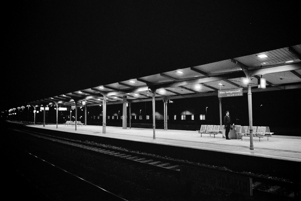 This image shows a dimly lit, empty train station platform at night. The roof is supported by evenly spaced columns, creating clean, geometric lines. One person stands alone near some benches under a sign that reads 'Wien Heiligenstadt.' The platform feels deserted, with no train in sight, and the scene is captured in black and white, emphasizing the stark contrast between the lights and the dark, empty space around it.