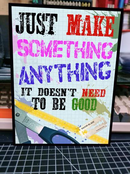 A colourful metal sign propped against a computer keyboard. The sign reads "just make something, anything, it doesn't need to be good" in a mixture of black, pink, red, purple, and green text. The bottom of the sign shows the handle of a pair of scissors, a tape measure, and a razor blade against a paint flecked background.