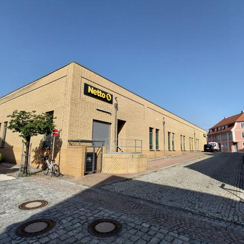 The light-coloured brick walls of a supermarket rise up under a bright blue sky. Three manhole covers interrupt the cobblestones of the surrounding street. A single tree breaks up the stone-heavy scenario.