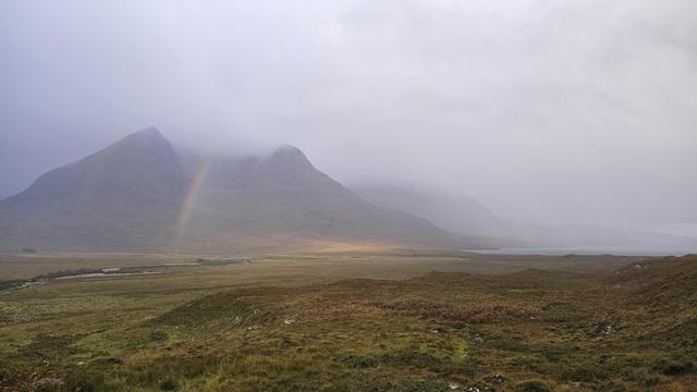 Misty mountain landscape with a faint rainbow in the Scottish Highlands. The serene scene features more brown than green fields in the foreground, with hills partially covered by clouds, creating a tranquil and atmospheric view. Ideal for nature enthusiasts and travel inspiration. A rainbow is partially visible beside a lake.