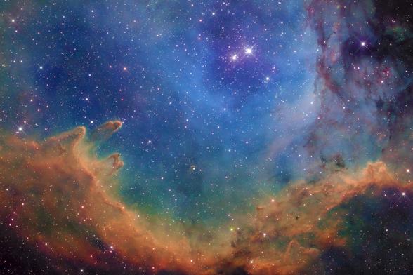 Astrophotography of a section of the Soul Nebula. In the bottom third of the image are bright orange and yellow gases forming a wave like structure with pillars extending into the rich blue background.