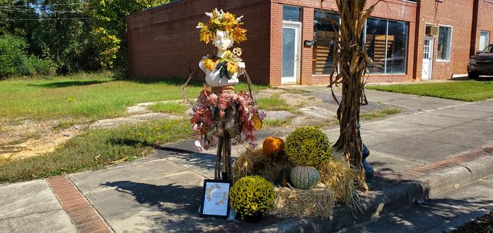 A half-mannequin dressed in a crown and necklace of autumnal flowers amd a skirt of red and white flowers situated near haybales and pumpkins all in front of an abandoned building. The sign in front reads "Fairy Witch."