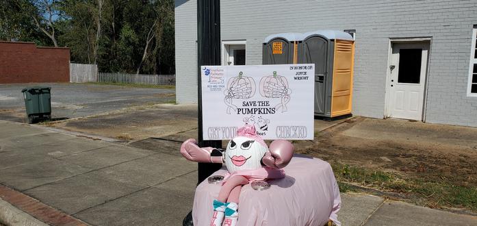 A white and pink pumpkin with big painted lips and eyes wearing boxing gloves and sitting atop a square hay bale with a sign behind it reading "Save the Pumpkins." It is part of the pink ribbon breast cancer research movement in the U.S.
