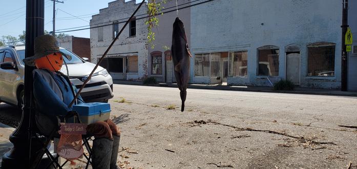 A jacko'lantern scarecrow sitting in a fabric folding chair with a catch of the day and a fish at the end of the line on a fishing rod all in front of a row of abandoned, decrepit buildings.