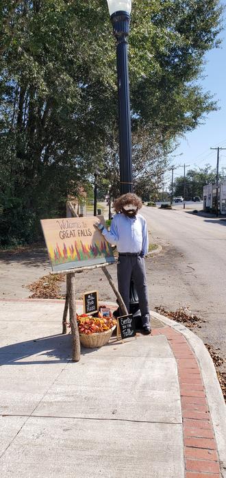 A Bob Ross style scarecrow next to a painted canvas with autumnal trees on it. The canvass reads "Welcomecto Great Falls." Beneath the canvas is a basket of small pumpkins.