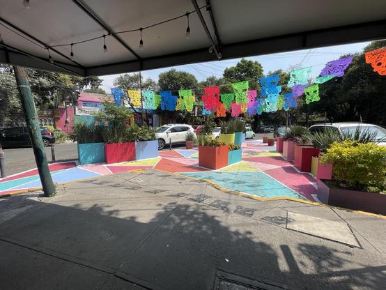 A segment of an intersection painted a multitude of bright colors, creating a plaza, with planters on the perimeter. There are colorful banner strung across the plaza. The sun is shining.