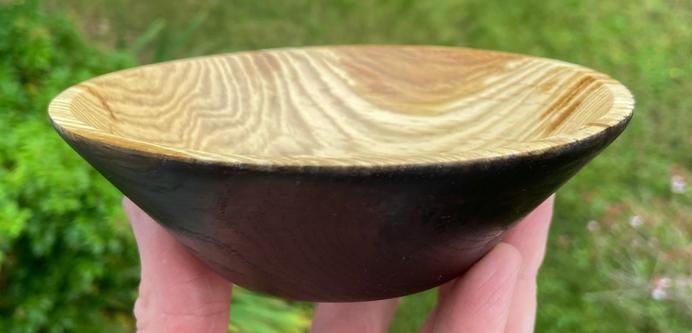 An oak bowl, resting on finger tips. The inside is brown and streaked with grain patterns. The outside is black with patterns from the grain as well.