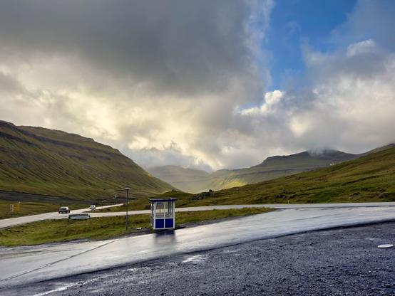 It just stopped raining, so the road is wet and the sky is about to open. A speck of blue is appearing between dark and yellow lit clouds. Treeless mountain slope limit the valley. A road crosses the lower part of the image, on the opposite side a bus stop cabin stands solitary.