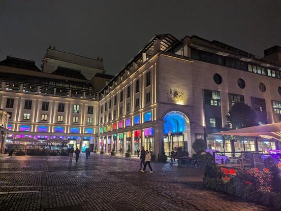 Night photo of a 3 storey Georgian white stone building with many arches which are lit underneath by different coloured lights.