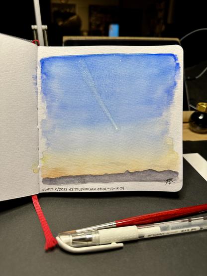 A watercolor on gray toned paper of the evening sky, scant clouds lit faintly with sunset, looking west across the Rogue Valley in southern Oregon. A range of mountains crosses the bottom of the scene. High above is a faint comet with a long tail stretching towards the upper left corner. A sprinkling of stars appear in the deepening sky. A paintbrush and white pen rest on a desk below the painting.