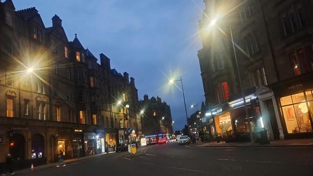 Looking east along Bruntsfield Place at night, lines of handsome Victorian stone tenements with lights of cafes, shops and restaurants on ground floors