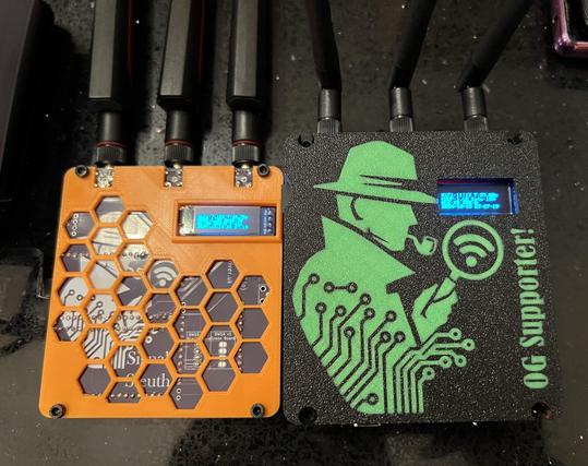 Two Signal Sleuth wardrivers side by side, one super thin, one challenging not with a battery pack built in. The left one is orange, the right one is black with a green Sherlock Holmes styled character in silhouette