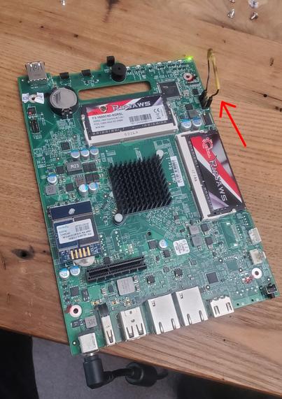 A small motherboard on a table with screws strewn about. There's a smudgy wire attached to a jumper on the motherboard. The board is plugged in, its green power LED lit up.