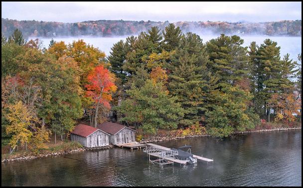 Two old boathouses along a relatively still late. Trees are turning yellow and red. In the background we steam or fog rising off the lake.