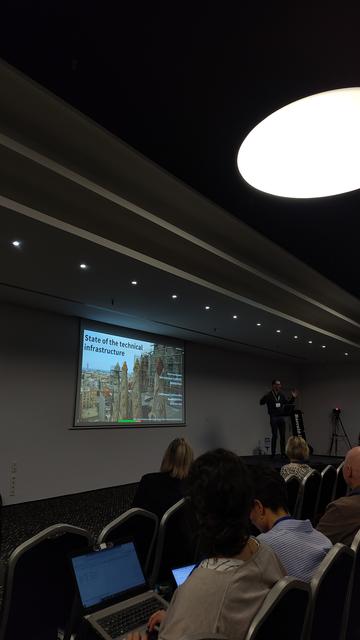 Conference room with a projected slide showing a picture of the Sagrada Familia and with the title "State of the technical infrastructure" superimposed, and a presenter on stage behind a lectern as well as a few members of the audience.