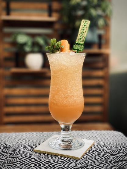 orange cocktail with mint sprig and orange peel garnish with a False Idol swizzle stick in a hurricane glass sitting on a white marble coaster with potted plants sitting on an acacia wood shelf in the background