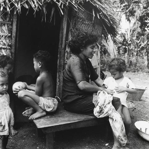 This black and white photograph depicts a scene from Longoteme, Tonga in 1963. A woman is sitting on a low wooden stool outdoors, engaged with one of her young children who she appears to be dressing or caring for using cloth draped over the child's torso. To her left sits another young boy squatting on the same bench holding a coconut close to his chest. Directly in front of them stands a young girl wearing a white dress looking towards something outside of the frame with an attentive expression, while to their right is yet another small child who seems slightly obscured and focused elsewhere.

Behind this group, there's partial visibility of what appears to be part of a wooden structure resembling a traditional fale (a type of house), constructed from wood and possibly thatch or other natural materials. The setting suggests an outdoor rural environment with lush greenery visible in the background hinting at tropical vegetation typical of Tongan landscapes.

The image captures a candid, everyday moment reflecting on the daily lives of these individuals within their community during that era.