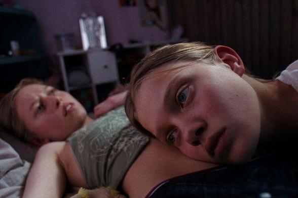 A girl lies on her back on a bed as a second girl listens to her stomach. Neither look happy. From Toxic