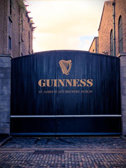 The image shows the iconic black gates of the Guinness Storehouse at St. James’s Gate Brewery in Dublin. The Guinness logo, featuring a golden harp, is displayed prominently on the gate, with the brewery name written below. The cobblestone street leads up to the entrance, framed by tall brick buildings on either side.
