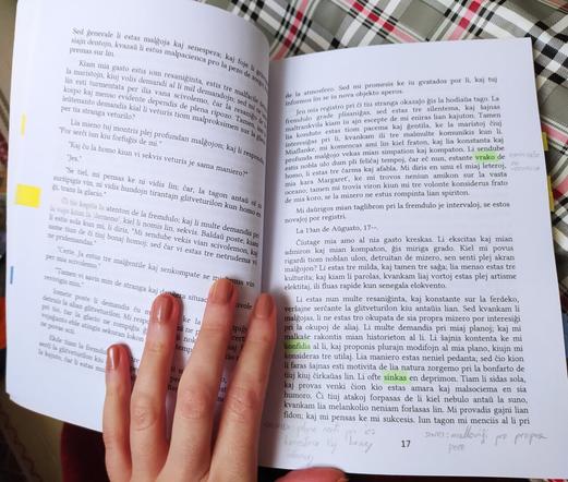 Mia mano tenas malfermita libron en Esperanto. La teksto estas markita tie-tie per verda surstrekilo kaj skribaĵoj el krajono. Ankaŭ kelkaj paĝoj estas markitaj per flavaj kaj bluaj legosignetoj.