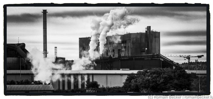 A black and white picture of an industrial site. Smoke comes up from the base of an industrial building. A tall chimney stands on the left side. No smoke is coming out of this chimney.  Dark clouds are hanging over the sky. The picture has a dark mood.