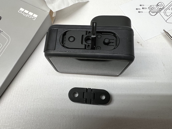 A GoPro HERO9 Black camera, upside down, with the folding fingers assembly from a new HERO13 camera installed instead of the regular HERO9 folding fingers (sitting below the camera on their own). The assembly fills the old folding fingers space almost perfectly.
