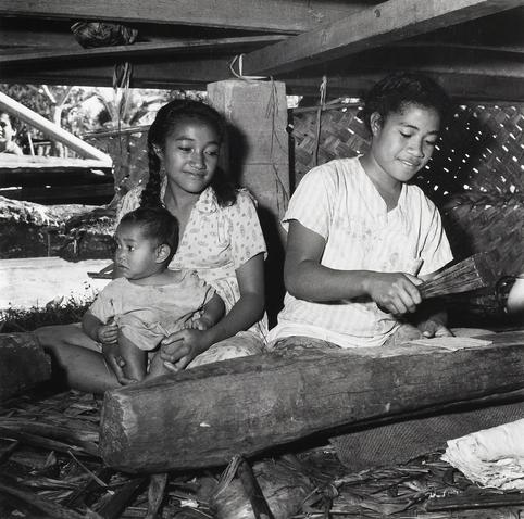 Two girls sit cross-legged on a woven mat, with one of them holding a young child in her lap. The girl to the right is using a small wooden mallet to beat paper mulberry bark on a large log as part of making ngatu (tapa cloth), while she sits under a traditional fale structure made from woven materials.