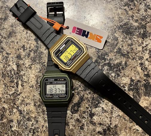 Two digital wristwatches are displayed side by side. The watch on the left is a classic Casio F91W in a dark green color with a rectangular face, showing the time 18:37:44. On the right is a gold-colored digital watch from Ali Express, with a similar retro design, also displaying the time as 18:37. Both watches have black rubber straps, and the Ali Express watch has a tag attached to it that reads "SKMEI." The watches are positioned against a plain background for comparison.