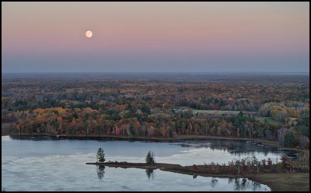 Aerial photo of a penisula sticking out into a calm lake just after sunset. The trees on the banks of the lake are showing their autumn colors and a full moon is visible in the sky.