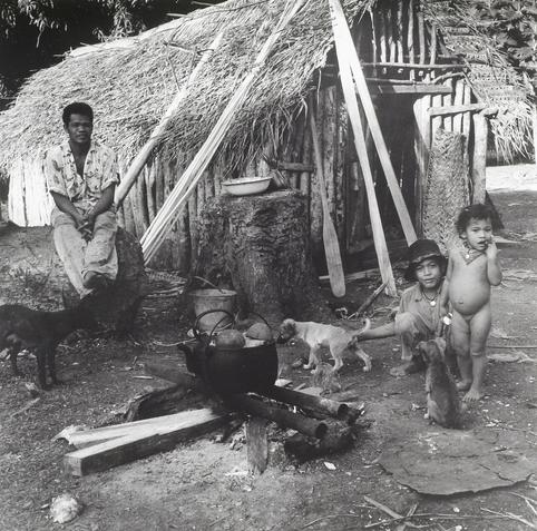 A black and white photo showing three individuals, including two children with various animals in a rural setting. One man sits on a tree stump to the left of another seated person who is behind him, while an unclothed child stands beside them interacting with other puppies that are present in this outdoor scene which seems to be from Longoteme, Tonga 1963.