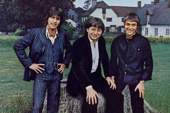 1977 

The image shows three men dressed in black suits standing next to each other on what appears to be a tree stump, with their hands casually placed on the trunk.
They are smiling and appear relaxed.
Tags: vintage, music group, 1970s fashion