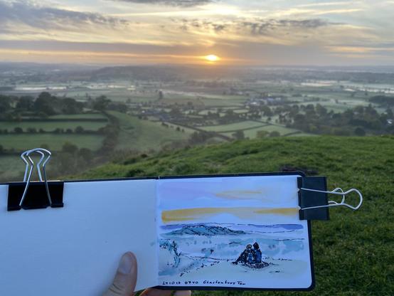 The sun rising above a green lamdscape with a coloured sketch of it in front.