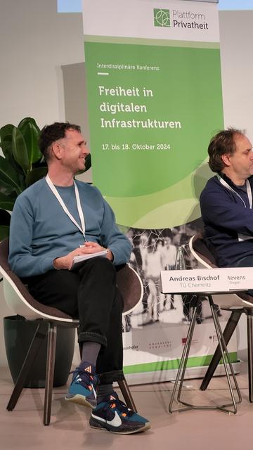 Links sitzt lächelnd Andreas Bischof, im Hintergrund ein Aufsteller mit dem Thema der Konferenz in Grün.