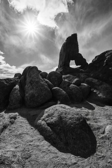 “Boot Arch” A black and white image featuring a prominent rock formation with an arch, surrounded by various large boulders. The sun shines brightly in the sky above, casting distinct shadows on the ground.