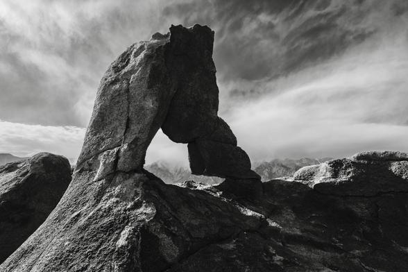 “Boot Arch” A dramatic black and white image of a large rock formation with an arch shape, set against a cloudy sky and distant mountains. The intricate textures of the stones are highlighted by shadows and light.