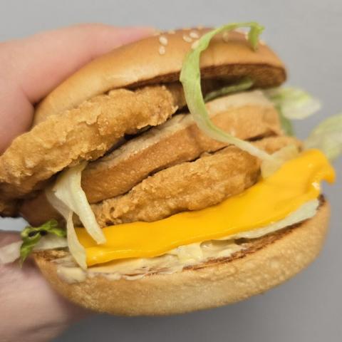 Photograph of a chicken sandwich held by a partially visible white person's hand against a light colored wall. It has two patties, a sesame seed bun, and lettuce / cheese are both visible. There's some kind of special sauce on it. It's a chicken Big Mac.