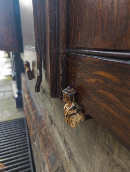 A wooden shutter is held open by moveable metal holders in the shape of a human head and shoulders. The nearer of the two holders is rusted and weathered.