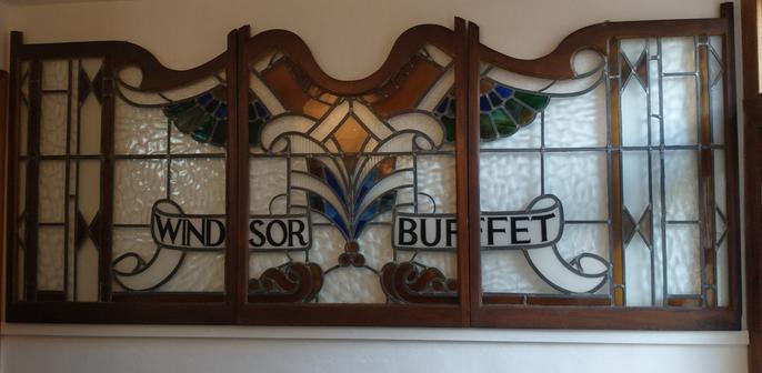 Three stained glass panels sit within an intricately carved wooden frame. The stained glass has a kind of coloured wing pattern and two white ribbons, with the words 'Windsor Buffet' written in them.