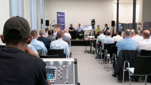 Das Bild zeigt die Aufnahme zum Podcast „Per Anhalter durch den Cyberraum“ mit der Podiumsdiskussion mit mehreren Teilnehmenden, die vor einem Publikum auf einer Bühne sitzen. Im Vordergrund sieht man ein Techniker, der ein Mischpult bedient und die Technik für die Aufnahme des Podcast überwacht. Die Veranstaltung scheint professionell organisiert zu sein, mit einem Kamerateam, das die Diskussion aufzeichnet. Im Hintergrund ist ein Banner zu erkennen, das auf die Veranstaltung oder den Veranstalter hinweist.
Foto: Cyberagentur