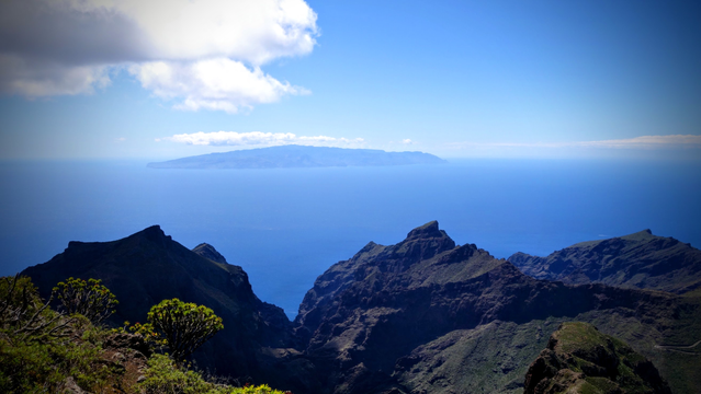Von einen Gipfel im Teno-Gebirge blickt man über grünlich bewachsene kantig martialisch aussehende steile Berge und Schluchten aufs diesige Meer. Am Horizont als dunkler Schatten eine Insel, die fast zu schweben scheint.