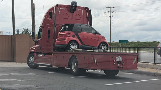 A semi tractor with a platform over the bread wheels instead of a trailer hitch parked at a McDs.  Sideways on the platform tucked up against the cab is a Smart Car.  Both are colored red.