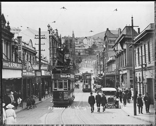 Black-and-white photo: Manners Street, Wellington. 1920s? Photographer unknown. Description: Looking west up Manners Street, Wellington showing buildings on right including a clothier, ladies’ tailor, signs for Billiards Upstairs (centre); Siegels for Keenest Value Costumier (centre); on left premises include: Cookham House with G.M. Fowlds Ltd. footwear; W. Campbell Sign Writers; tower of St Mary of the Angels (left rear) houses on hill (rear); pedestrians on footpaths [BrE pavements, AmE sidewalks] and crossing road – there are no obvious pedestrian crossings or ‘safety zones’; cars (right foreground to rear and left centre); two double-decker trams (left foreground and centre) with a single decker tram behind (largely obscured rear). Citation: Auckland Libraries Heritage Collections 35-R1784. https://kura.aucklandlibraries.govt.nz/digital/collection/photos/id/274853