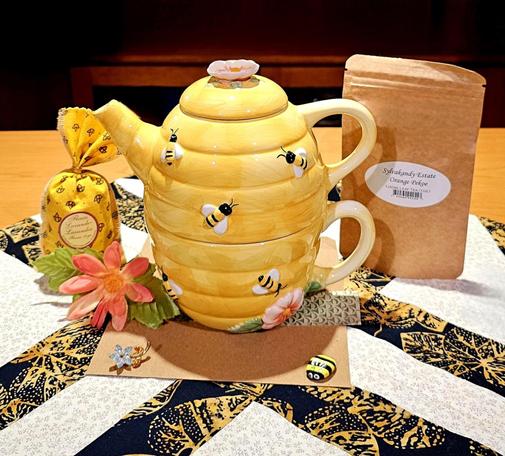 A bee-themed tea tableau in yellow, white, gold, and black. At center is a tall tea-for-one set shaped like a beehive and decorated with bees & garnished with peach- colored blossoms. To one side is a small yellow & black fabric bag filled with lavender,  to the other is a brown kraftpaper packet of Orange Pekoe Cut black tea from Sylvakandy Estate. At the base of the tea set are a pebble handpainted to look like a bee, and a tiny, dainty, jeweled brooch in the shape of a flower. Underneath everything is a quilting square in bold, long patchwork strips of black and gold batik interspersed with white- on- white strips.